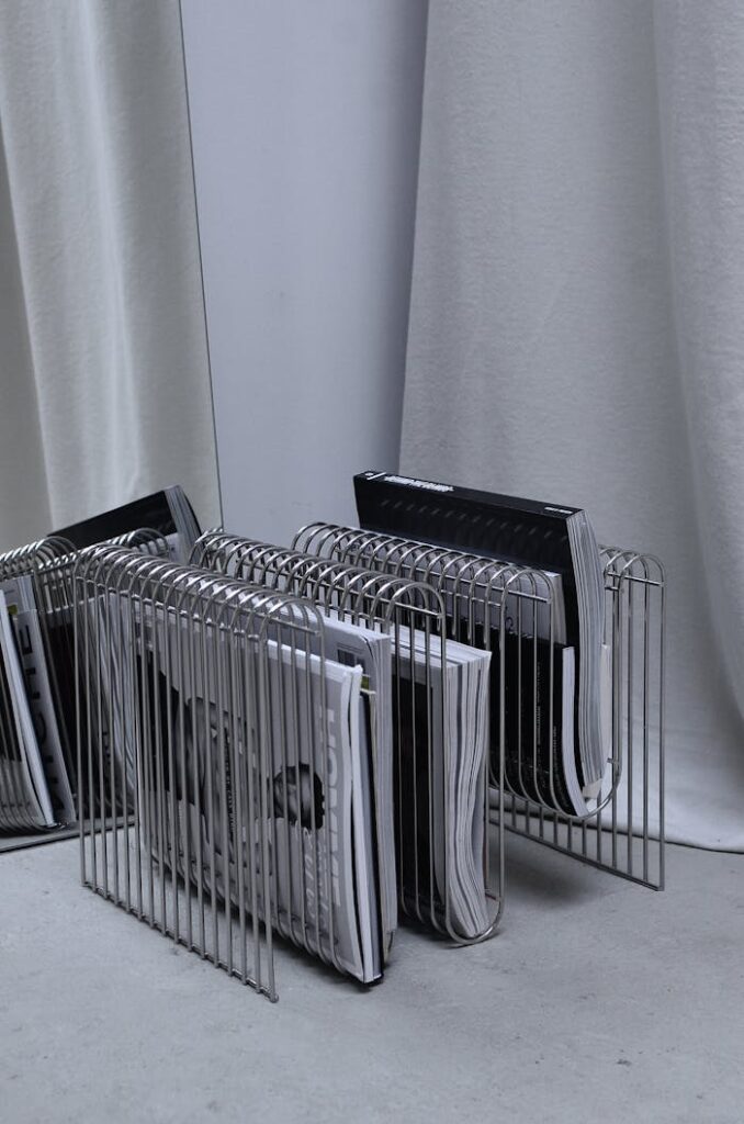 Collection of modern fashion magazines on metal rack placed on floor near mirror and curtain in room corner